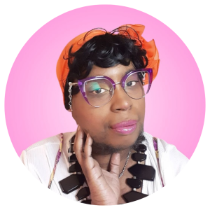 A Black feminine person with oversized purple cat eye glasses, big black chunky necklace, a curly pixie cut wig, and a orange flower hat. A Photo of Gigi Kiersten